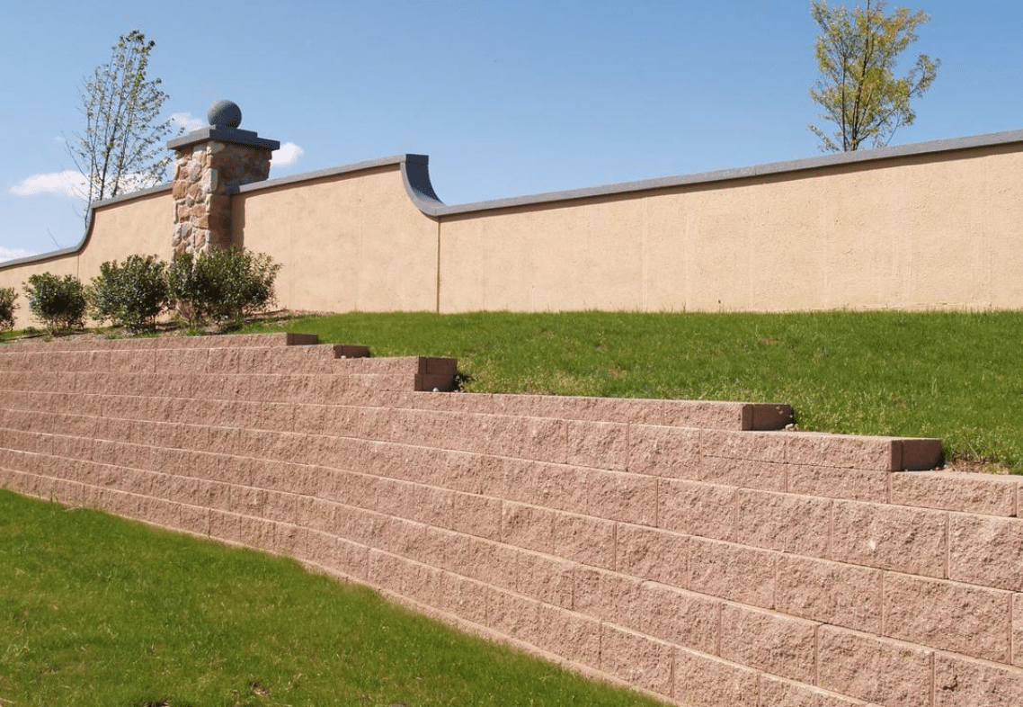 A beige concrete retaining wall with multiple levels supports a grass slope. There is a stone pillar and a beige, stucco wall with decorative accents at the top. Trees are visible in the background.